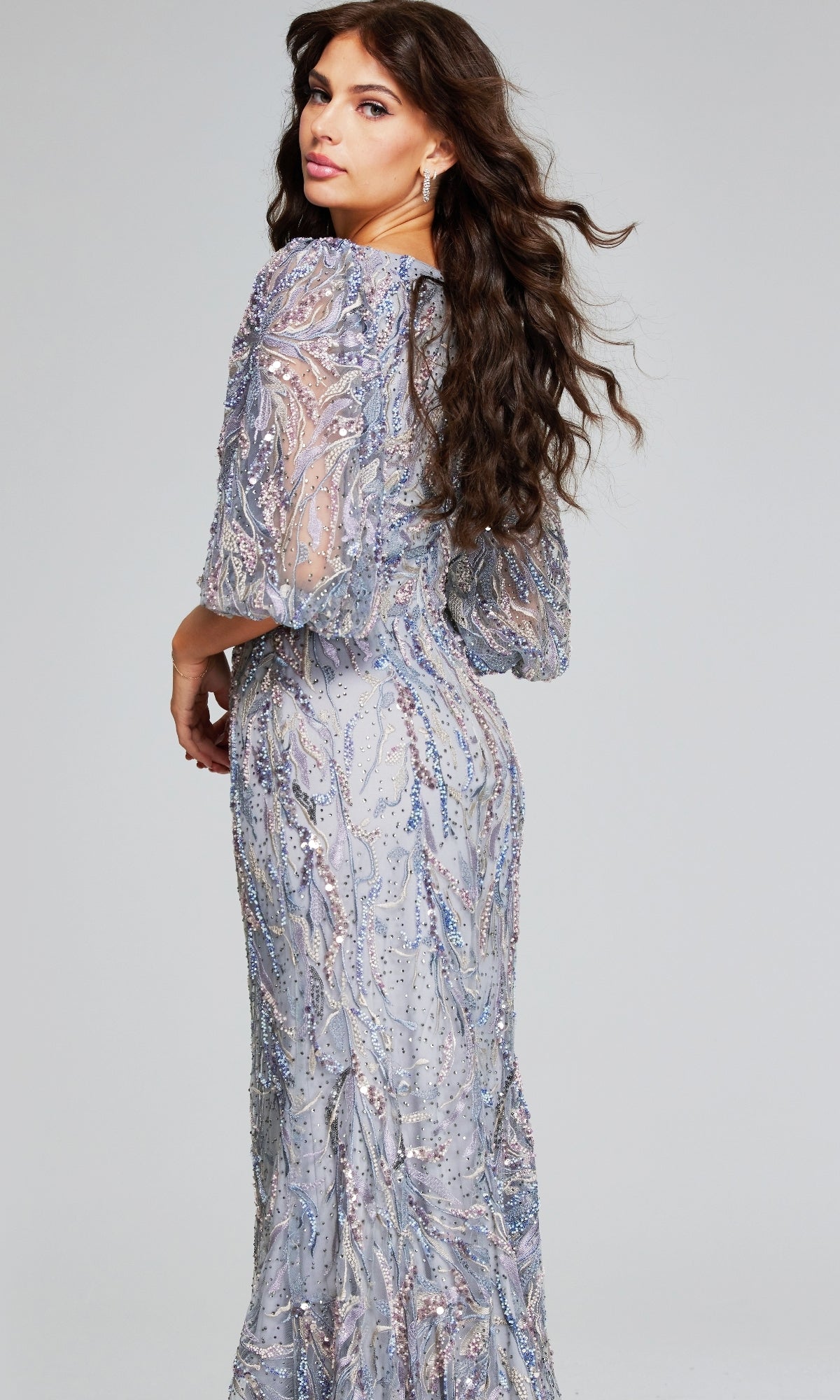 Woman with long brown hair wears the Jovani 38110 long grey formal dress, featuring sparkly embellishments, as she stands against a plain background looking over her shoulder.